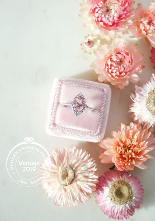 Vaaleanpunainen halo-mallinen timanttisormus sormusrasiassa ympärillään erivärisiä kukkia