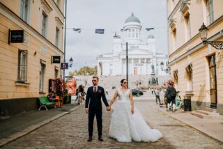 Hääpari aurinkolaseissa seisomassa käsikkäin keskellä kaupunkia ja mukulakivikatua taustallaan Helsingin Tuomiokirkko.