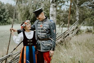 Hääpari vierekkäin pellon laidalla nojaamassa aitaan ja pukeutuneena kansallispukuihin ja kukkaseppeleeseen
