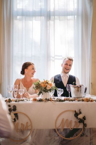 Hääpari nauraa hääparin pöydässä, jota koristaa hääkimppu ja Mr. ja MRs. -kyltit